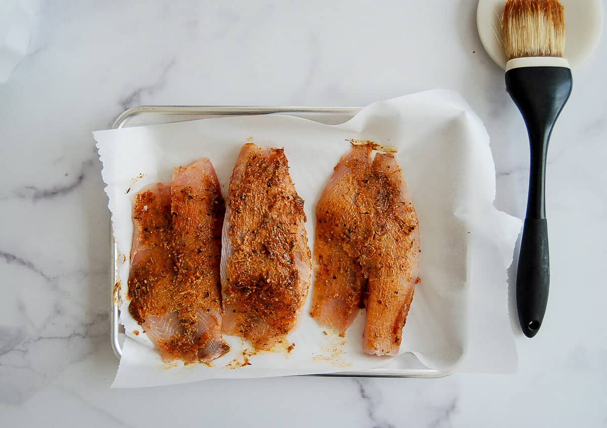 Three raw tilapia fillets coated with spices are arranged on a parchment-lined baking sheet. A basting brush rests on a small plate to the right, all placed on a white marble surface.