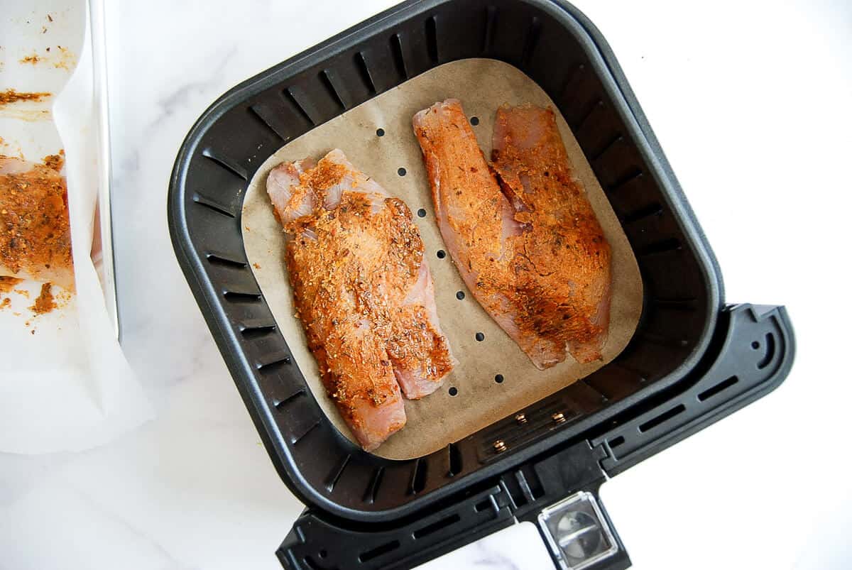 Two pieces of seasoned tilapia fillets are placed on parchment paper in an air fryer basket, ready to be cooked.