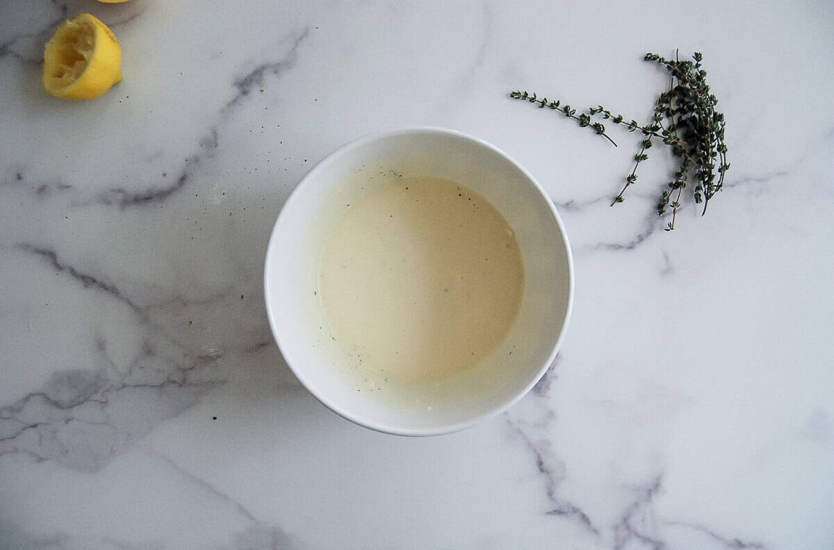 A white bowl containing a garlic aioli sauce sits on a marble surface, with a squeezed lemon half and fresh thyme sprigs nearby.