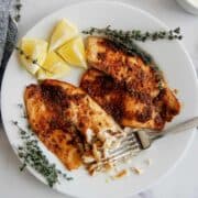 A white plate with two seasoned, air fryer tilapia fillets, garnished with sprigs of thyme, lemon wedges, and a fork flaking a piece of fish.