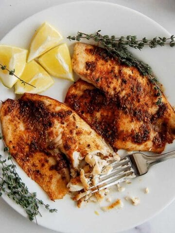 A white plate with two seasoned, air fryer tilapia fillets, garnished with sprigs of thyme, lemon wedges, and a fork flaking a piece of fish.