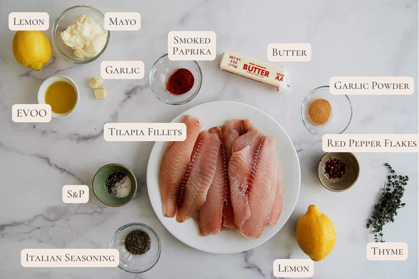 A plate of raw tilapia fillets surrounded by small bowls and ingredients, including lemon, mayo, garlic, smoked paprika, butter, garlic powder, red pepper flakes, thyme, Italian seasoning, salt, pepper, and olive oil.