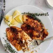 A white plate with two pieces of seasoned, cooked fish fillets, garnished with fresh thyme and lemon wedges. A fork is flaking a piece of the fish. A small bowl of white sauce is in the background.