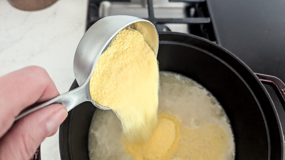 A hand pours yellow cornmeal from a metal measuring cup into a black pot of simmering liquid on a stovetop.