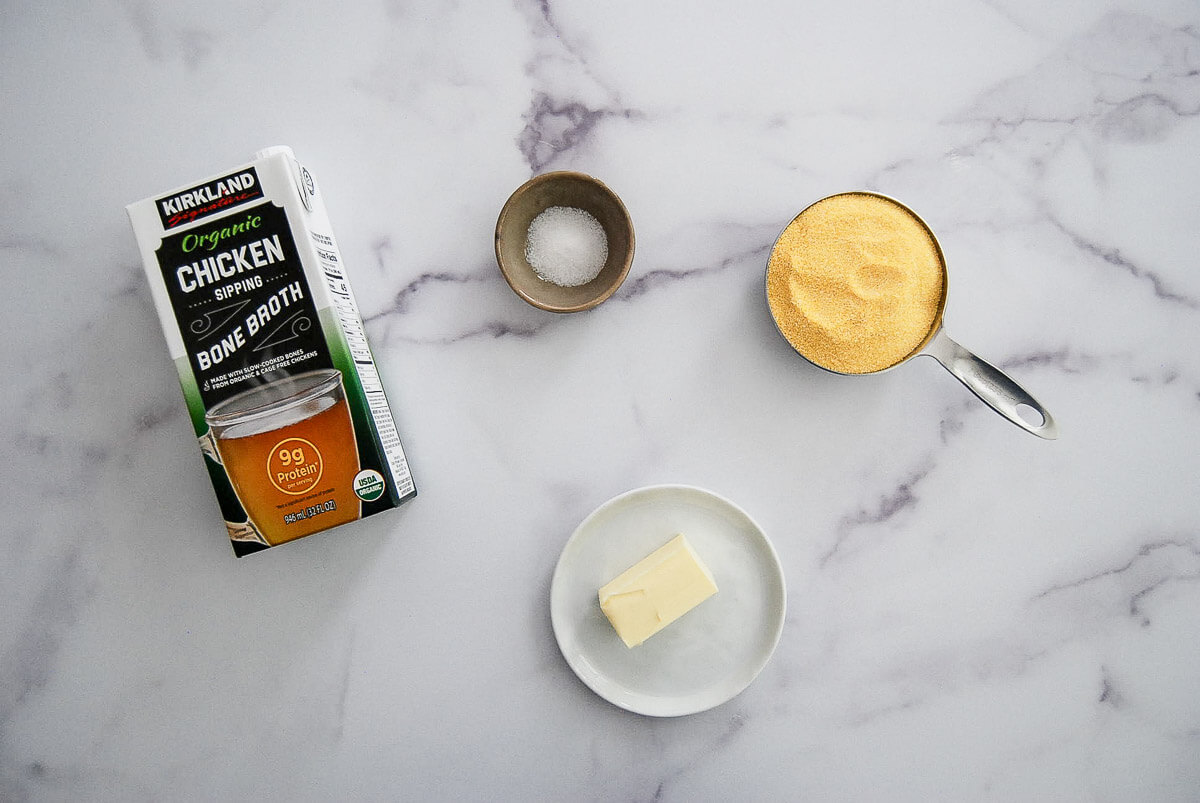 A carton of Kirkland chicken bone broth, a small bowl of salt, a cup of yellow cornmeal, and a plate with a pat of butter are arranged on a marble surface.
