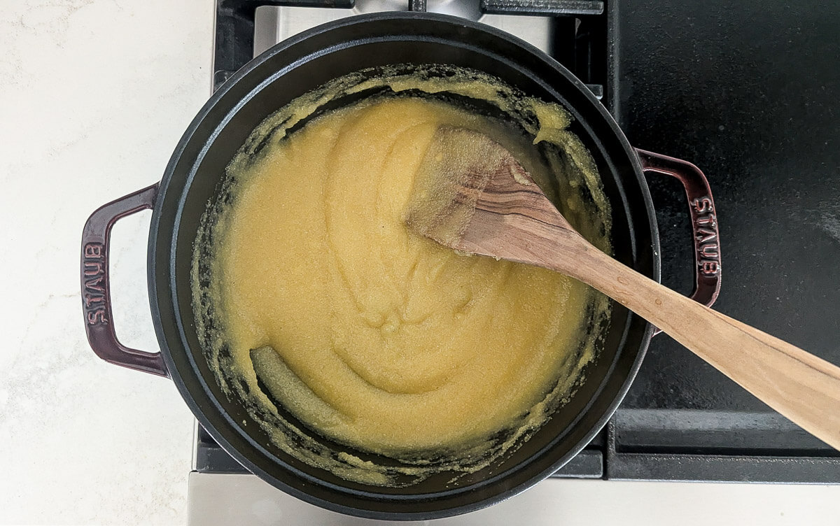 A wooden spoon stirs a polenta mixture in a black pot on a stovetop.
