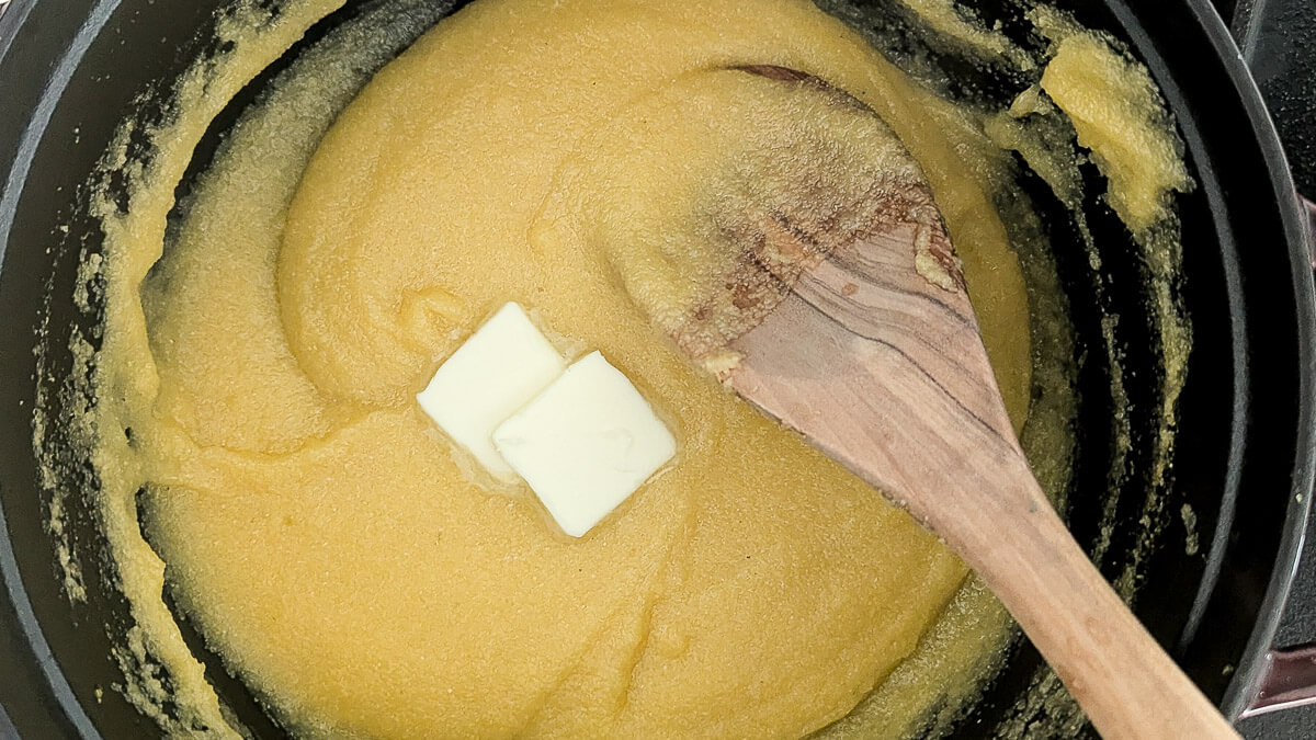 A pot of thick yellow polenta being stirred with a wooden spoon, with two small cubes of butter melting on top.