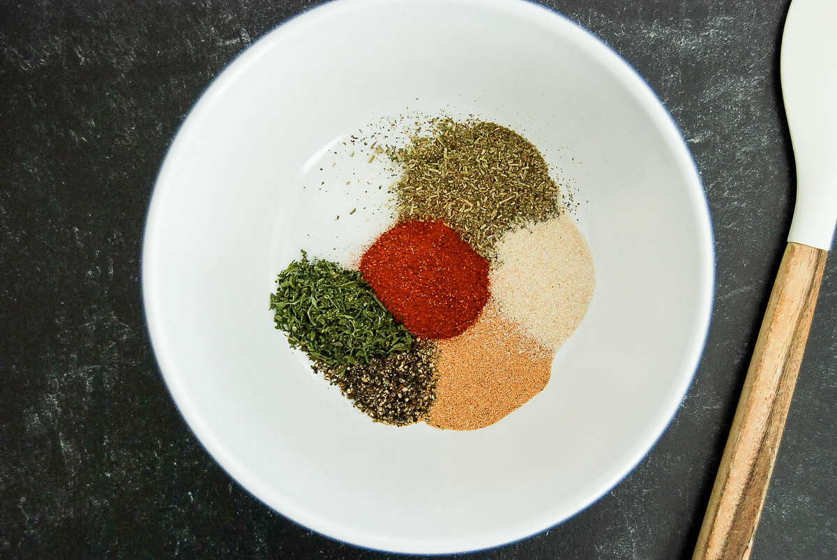 A white bowl contains piles of dried herbs and spices, including green herbs, black pepper, paprika, and granulated powders. A white spoon with a wooden handle is beside the bowl on a dark surface.