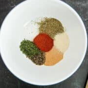A white bowl contains small piles of various dried herbs and spices, for a meatloaf seasoning mix, including green, red, brown, and off-white powders, arranged separately on a dark surface.