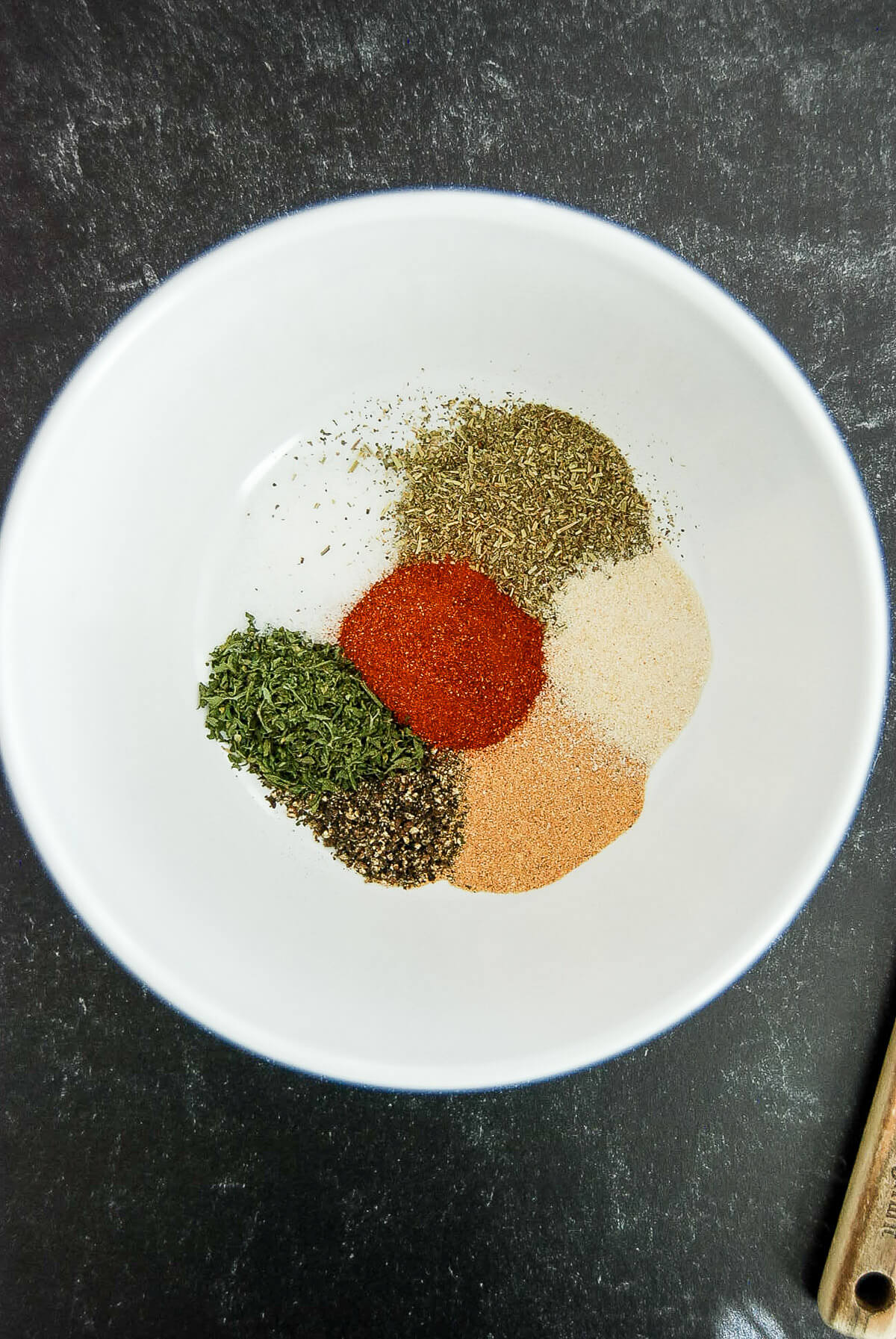 A white bowl contains small piles of various dried herbs and spices, for a meatloaf seasoning mix, including green, red, brown, and off-white powders, arranged separately on a dark surface.