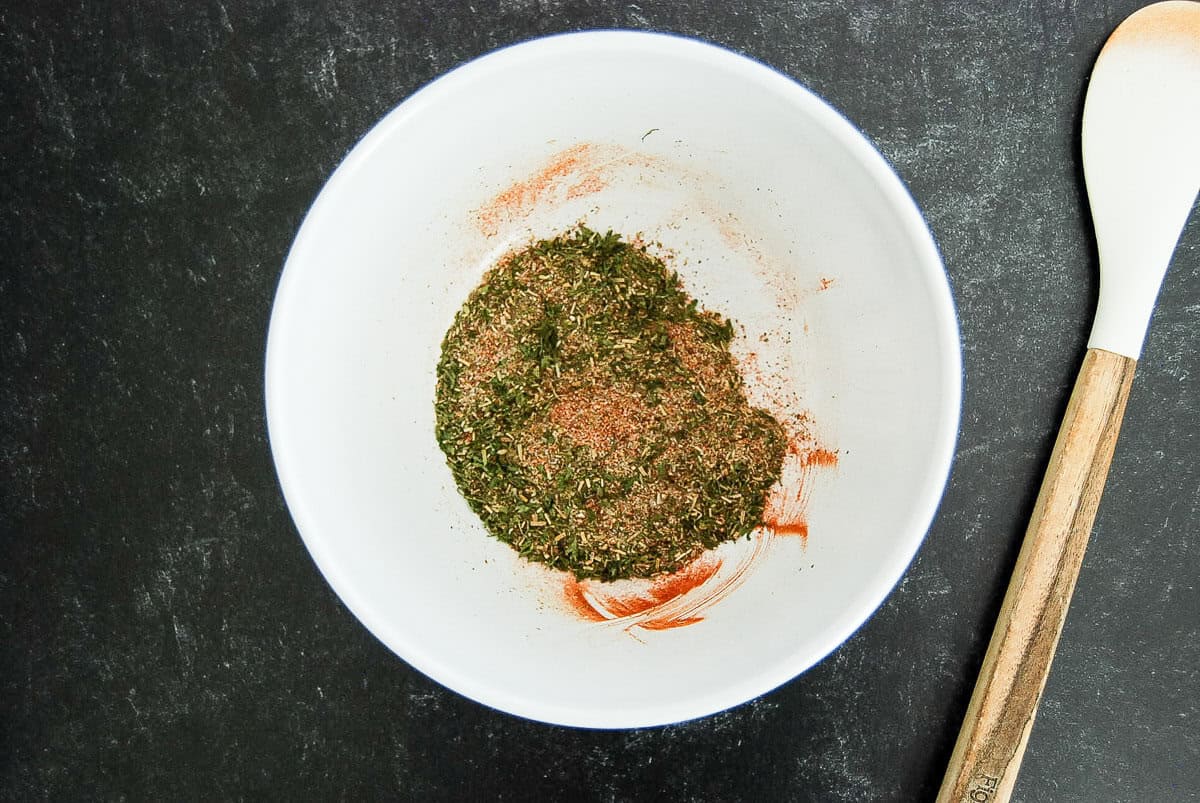 A white bowl with mixed dried herbs and spices for a meatloaf seasoning mix, on a dark surface, next to a wooden spoon with a white handle.