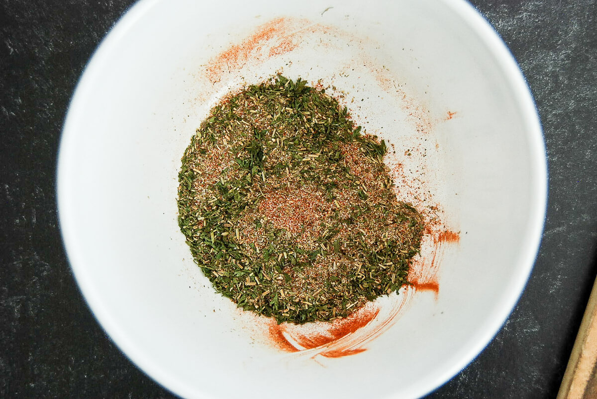 A white bowl containing a blend of green dried herbs and red spices, for a meatloaf seasoning mix, sitting on a dark countertop. Some of the spice mixture is spread along the sides of the bowl.