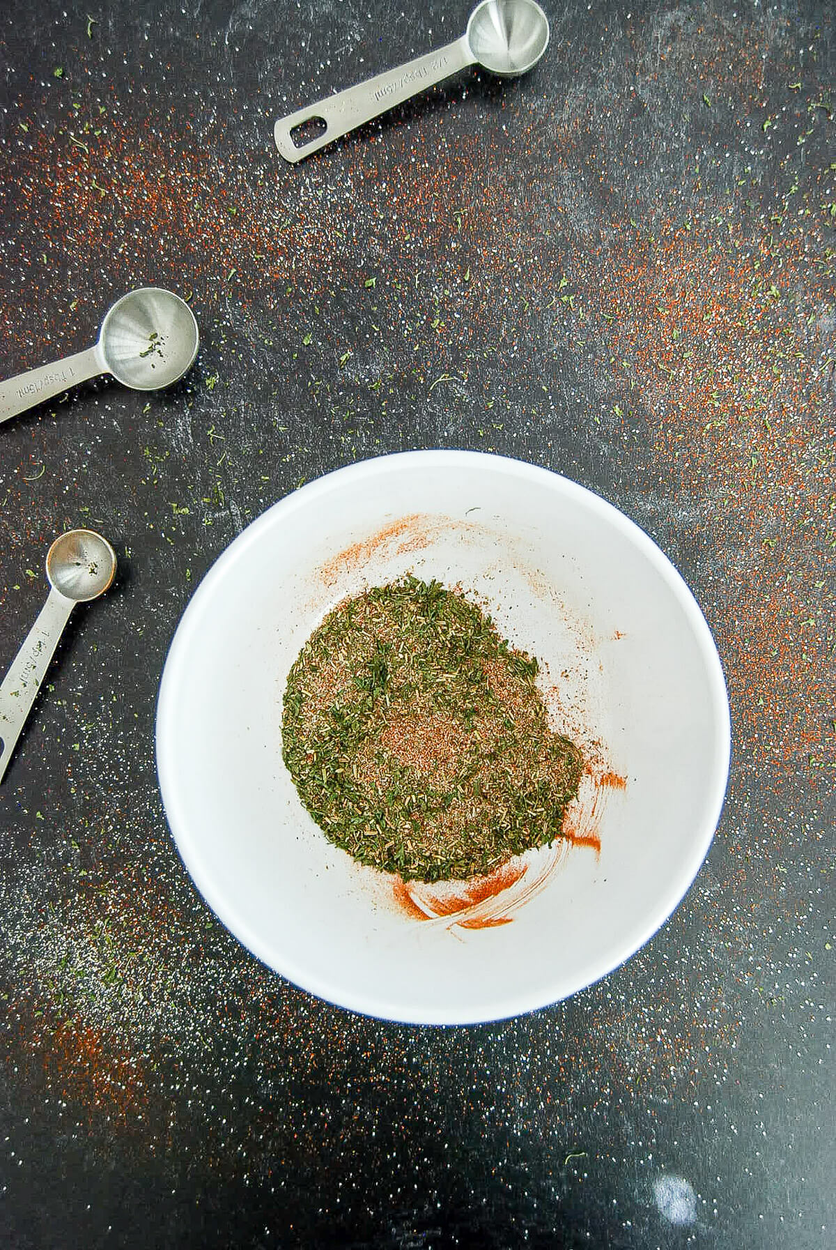 A white bowl with mixed dry spices for a meatloaf seasoning blend, sits on a dark surface, surrounded by scattered spices and three metal measuring spoons.