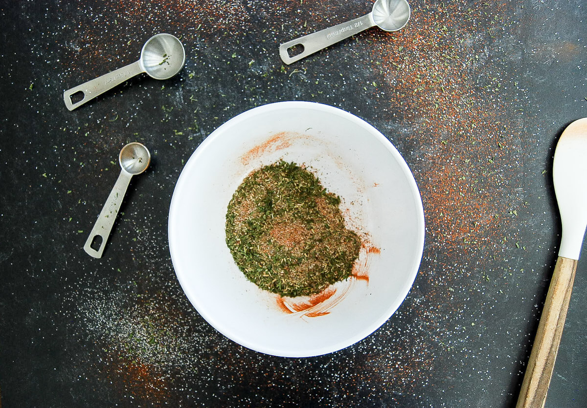 A white bowl with mixed dried herbs and spices sits on a dark surface, surrounded by four metal measuring spoons and a wooden spoon. Some seasoning is scattered on the tabletop.