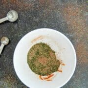 A white bowl with dried herbs and spices inside, placed on a dark speckled surface with some seasoning scattered around. Metal measuring spoons lie nearby.