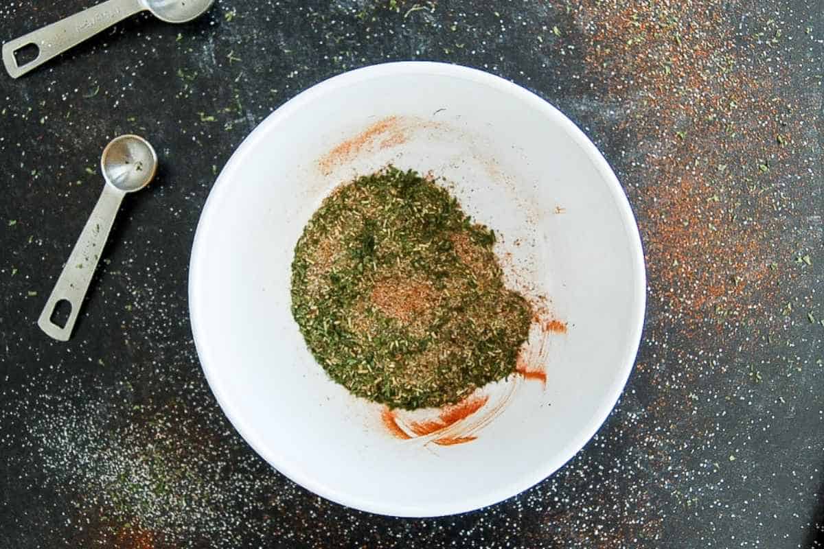 A white bowl containing a mixture of dried herbs and spices sits on a dark speckled surface. Two metal measuring spoons and scattered spices are visible around the bowl.
