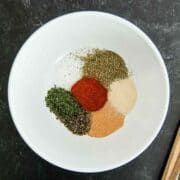 A white bowl containing small piles of dried herbs and spices, including paprika, black pepper, parsley, garlic powder, onion powder, and Italian seasoning, on a dark surface.