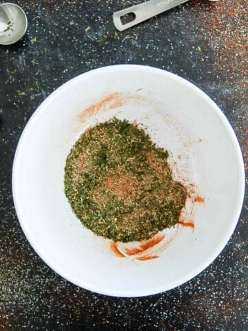 A white bowl filled with a mix of dried herbs and spices sits on a dark, speckled countertop, surrounded by three metal measuring spoons and scattered seasoning.