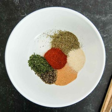 A white bowl containing small piles of dried herbs and spices, including paprika, black pepper, parsley, garlic powder, onion powder, and Italian seasoning, on a dark surface.