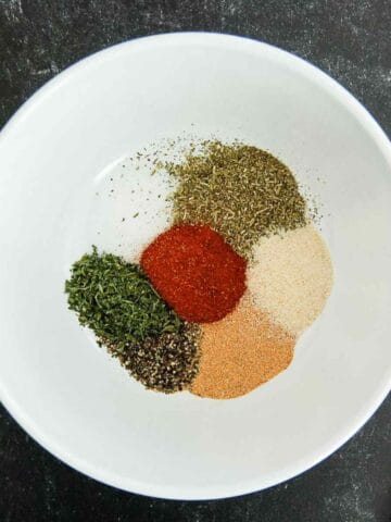 A white bowl containing small piles of dried herbs and spices, including paprika, black pepper, parsley, garlic powder, onion powder, and Italian seasoning, on a dark surface.