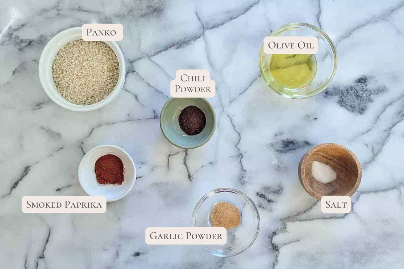 Six ingredients on a marble surface, each labeled: panko breadcrumbs, chili powder, olive oil, smoked paprika, garlic powder, and salt, all in small glass or wooden bowls.