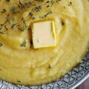 A close-up of creamy yellow polenta in a patterned bowl, topped with a pat of butter and garnished with fresh thyme sprigs.