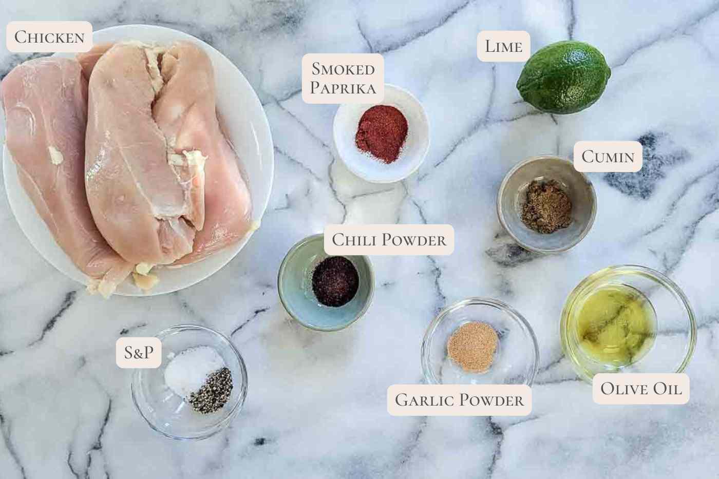 A marble surface with labeled ingredients for a chicken recipe: raw chicken breasts, salt and pepper, smoked paprika, chili powder, garlic powder, cumin, lime, and olive oil in small bowls.