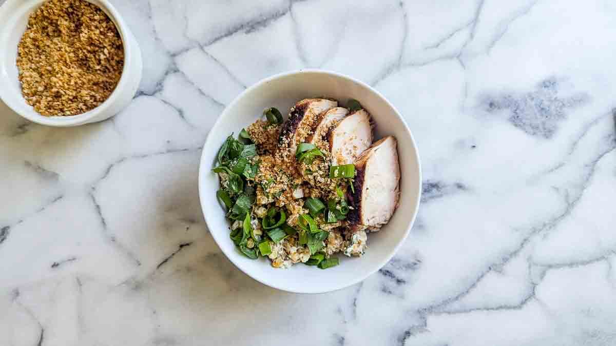 A white bowl on a marble surface contains rice, sliced chicken breast, mexican street corn, and green onions.