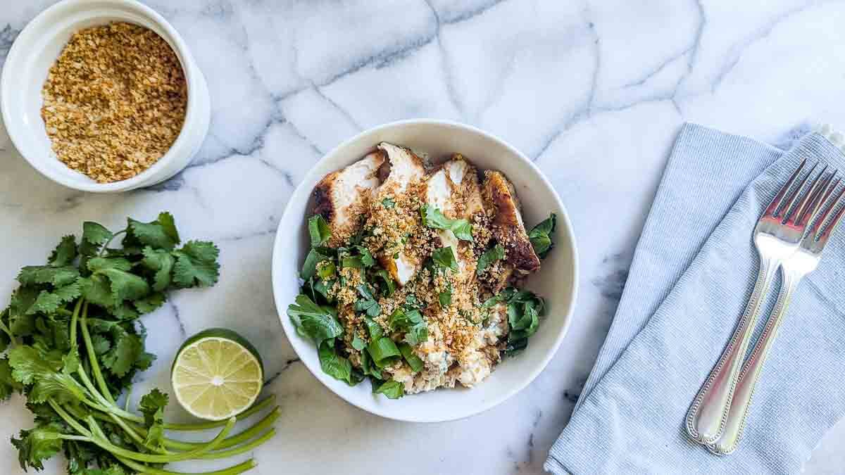 A white bowl on a marble surface contains rice, sliced chicken breast, mexican street corn, scallions, cilantro, and seasoned panko breadcrumbs.