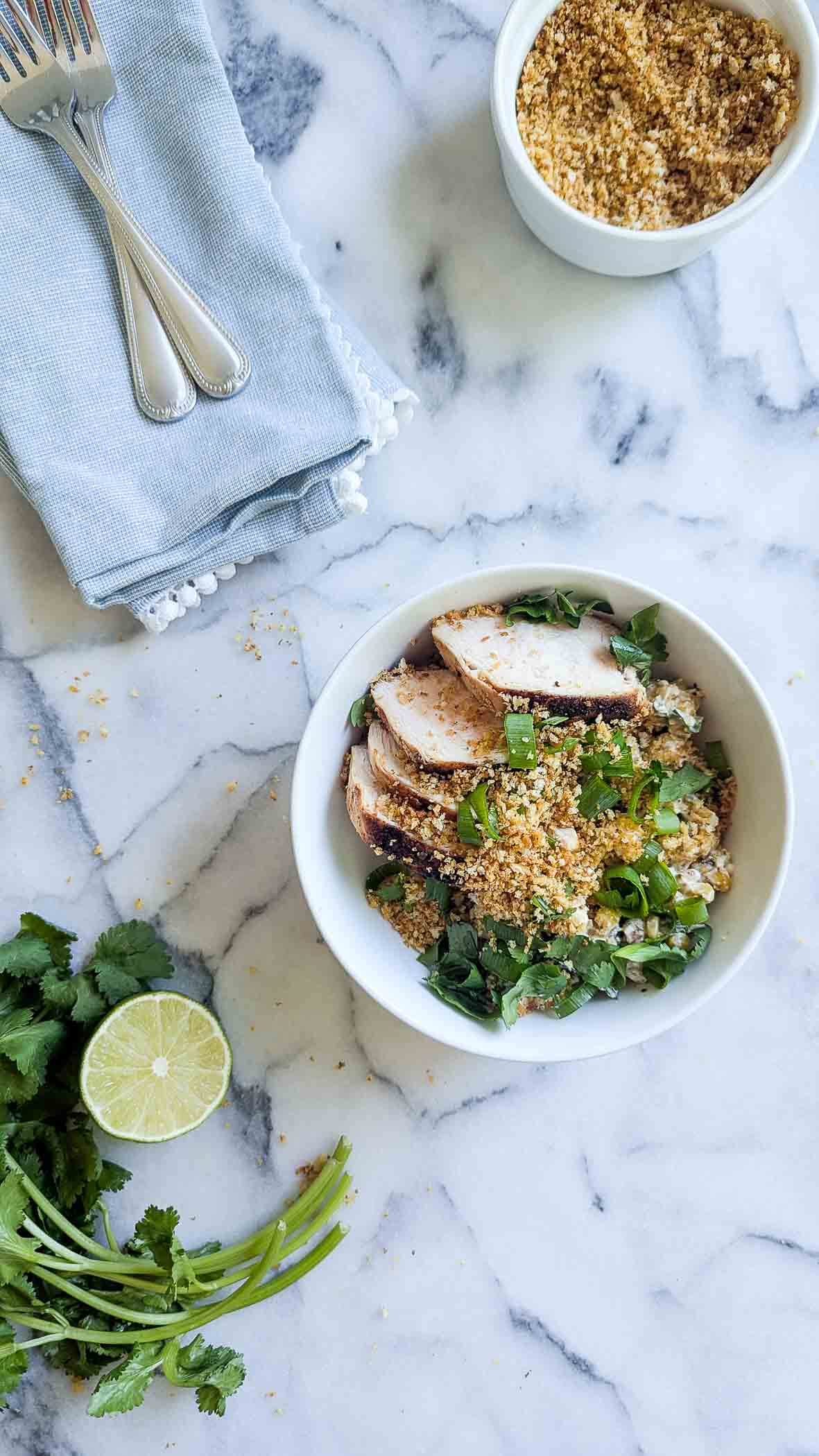 a street corn chicken and rice bowl on a marlble countertop with cilantro and lime on the side.