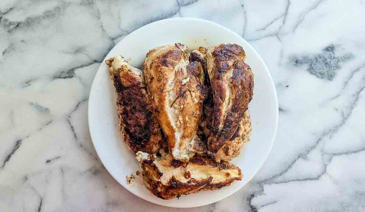 A white plate with four pieces of seasoned, cooked chicken breast sits on a marble surface. The chicken is browned and has visible grill or sear marks.