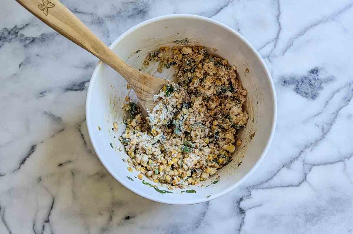 A white bowl with a wooden spoon contains a mixture of mexican street corn.