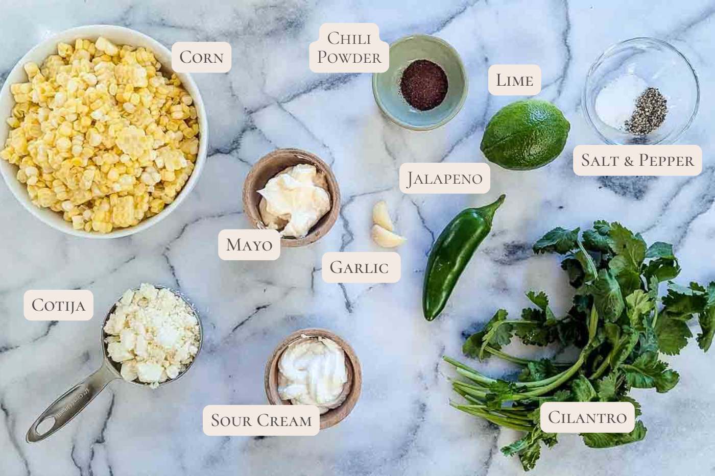 Ingredients for Mexican street corn spread out on a marble surface, labeled: corn, chili powder, lime, salt & pepper, jalapeno, cilantro, cotija cheese, sour cream, mayo, and garlic.