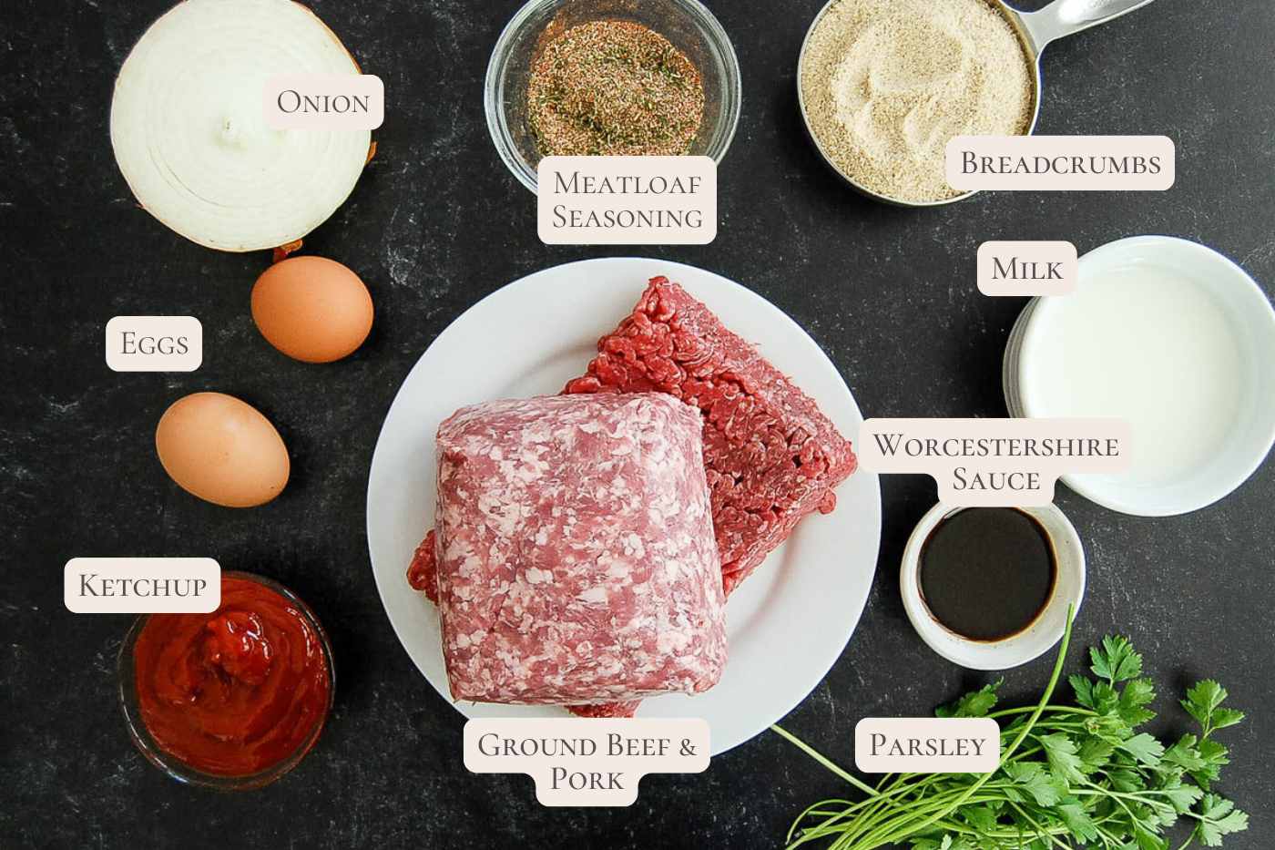 Top-down view of meatloaf ingredients: ground beef and pork, onion, eggs, ketchup, meatloaf seasoning, breadcrumbs, milk, Worcestershire sauce, and fresh parsley arranged on a dark background.