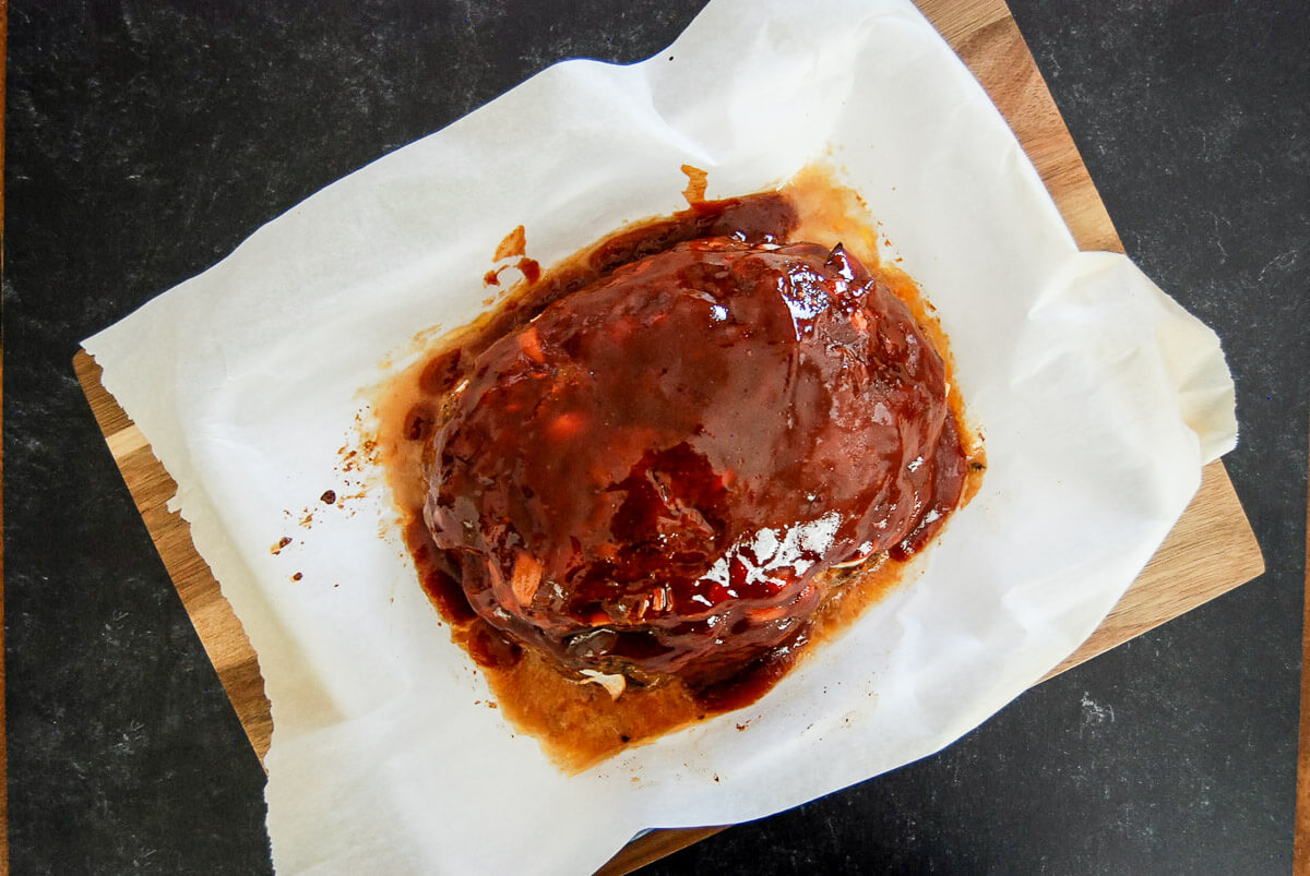 A glazed traditional meatloaf sits on a piece of white parchment paper atop a wooden cutting board, with meatloaf sauce pooled around it, on a dark countertop.