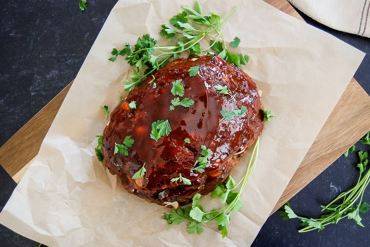 A glazed meatloaf garnished with fresh parsley sits on brown parchment paper atop a wooden cutting board, with additional parsley scattered around.