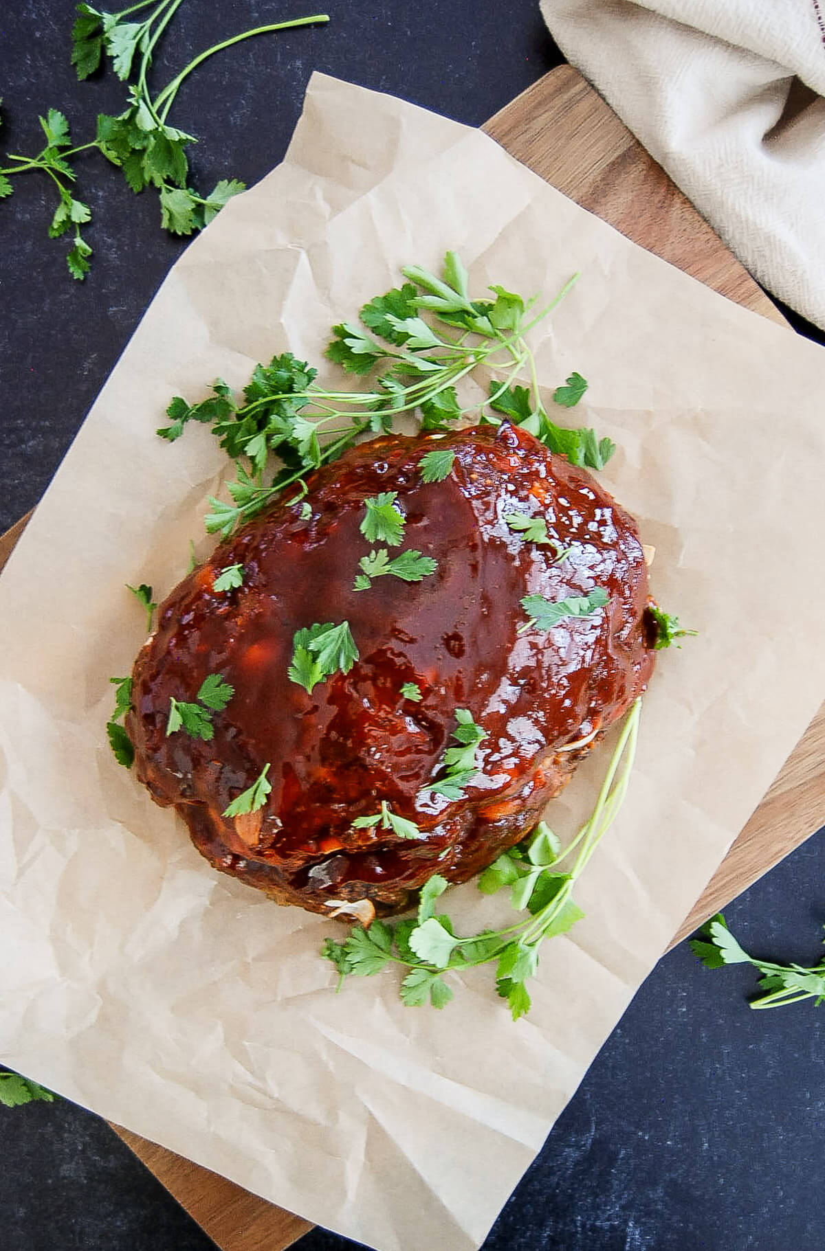 A classic glazed meatloaf garnished with fresh parsley sits on parchment paper atop a wooden cutting board, with additional parsley scattered around.