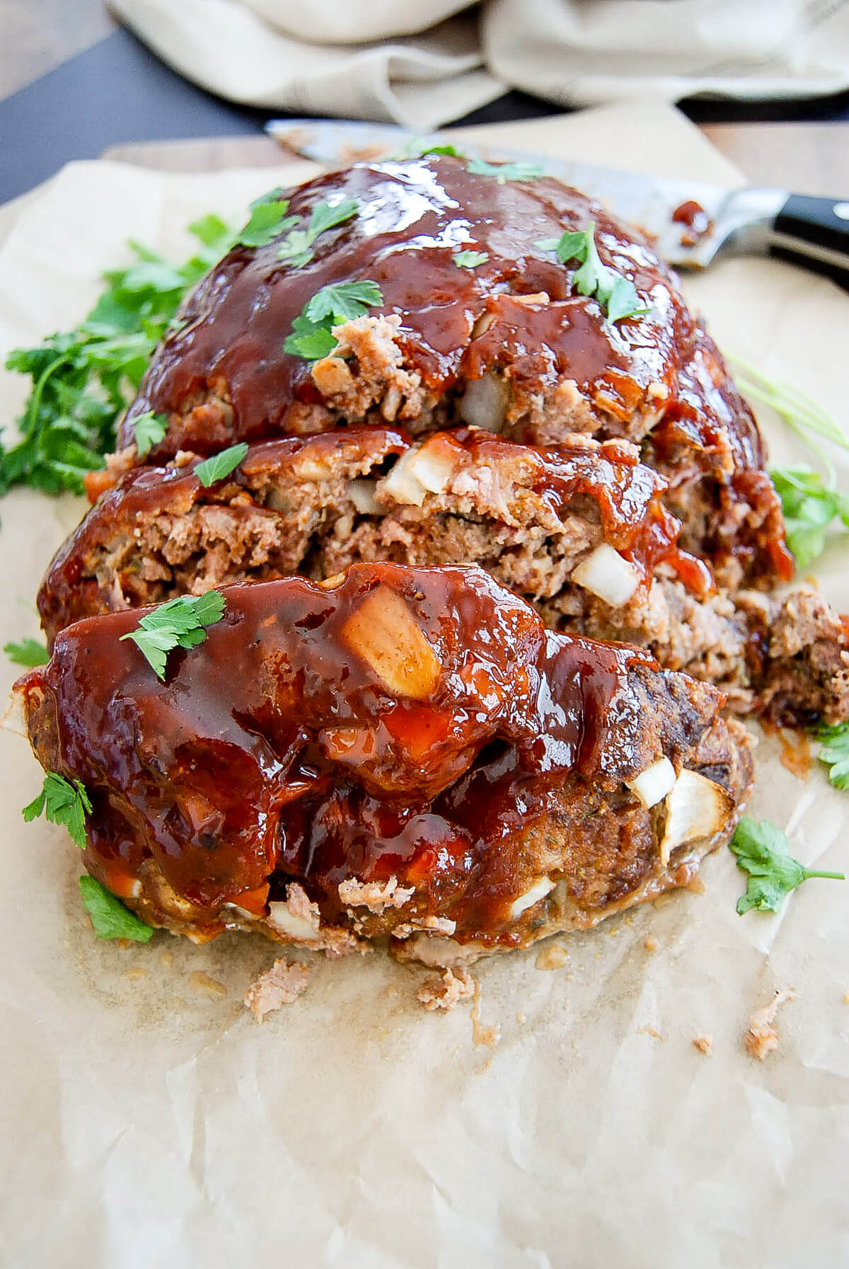 A sliced meatloaf topped with glossy meatloaf glaze and garnished with fresh herbs, sitting on parchment paper with some greens in the background.