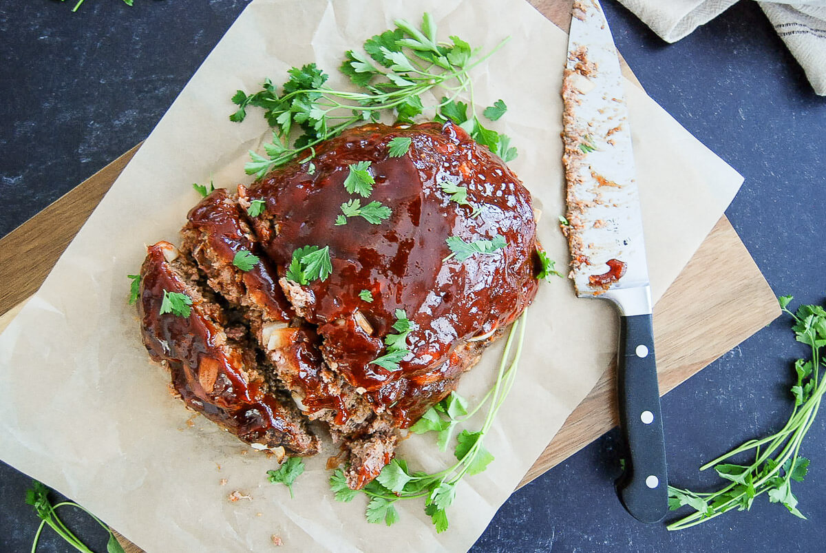 A glazed meatloaf garnished with fresh parsley sits on parchment paper beside a knife, with extra parsley scattered around on a dark surface.