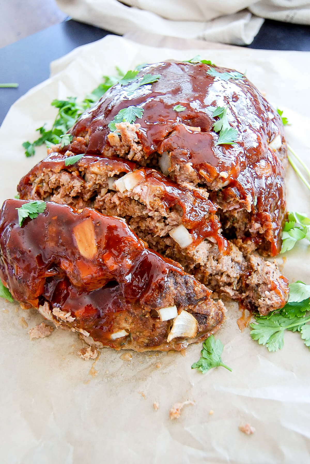 A glazed meatloaf topped with meatloaf sauce and garnished with fresh parsley sits on parchment paper, with a slice cut to reveal chopped onions inside.