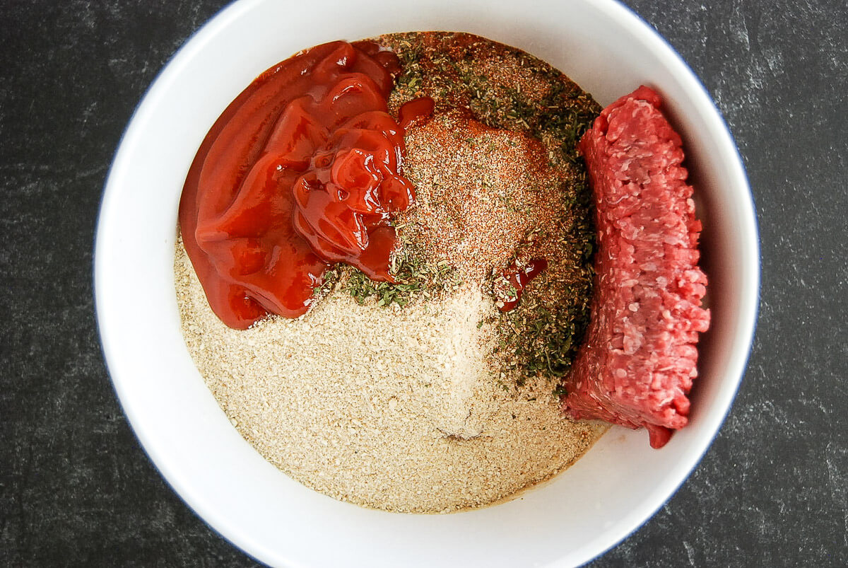 A white bowl contains ground beef, ground pork, ketchup, dried herbs, breadcrumbs, and spices on a dark countertop, with the ingredients separated and not yet mixed.