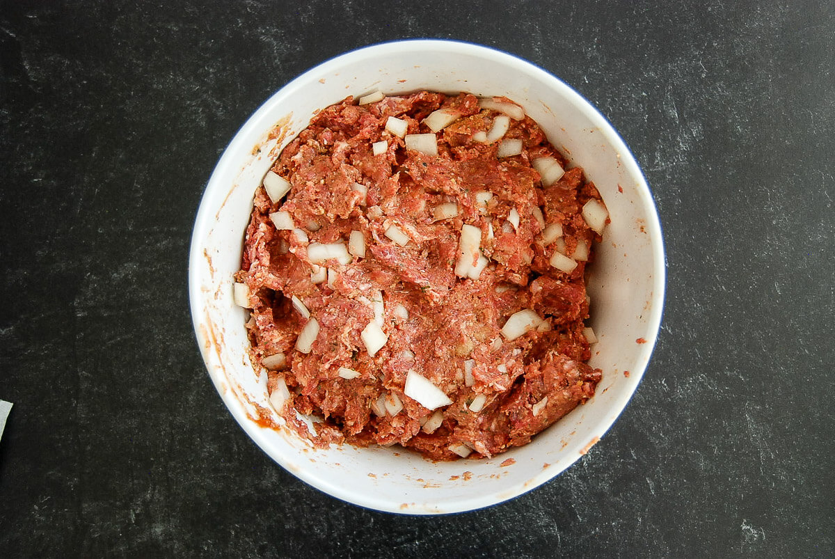 A white bowl filled with a mixture of ground beef and pork, chopped onions, and spices sits on a dark countertop.