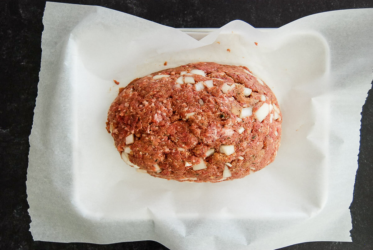 A raw meatloaf mixture with chopped onions is formed into an oval shape and placed on a sheet of parchment paper in a baking dish, ready to be cooked.
