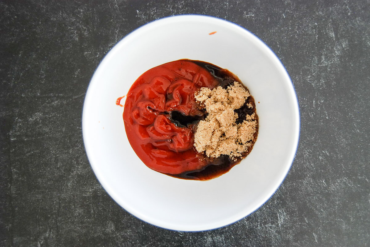 A white bowl containing ketchup, Worcestershire sauce, and a heap of brown sugar on a dark countertop.