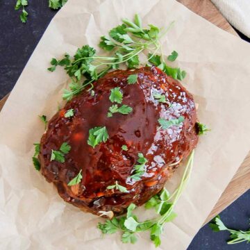 A glazed traditional meatloaf rests on brown parchment paper, garnished with fresh parsley sprigs and chopped parsley on top. The setup is on a wooden board.