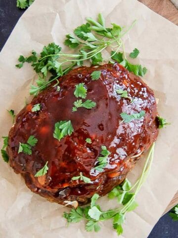 A glazed traditional meatloaf rests on brown parchment paper, garnished with fresh parsley sprigs and chopped parsley on top. The setup is on a wooden board.