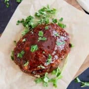 A glazed classic meatloaf topped with fresh parsley sits on parchment paper on a wooden board, with sprigs of parsley scattered around.