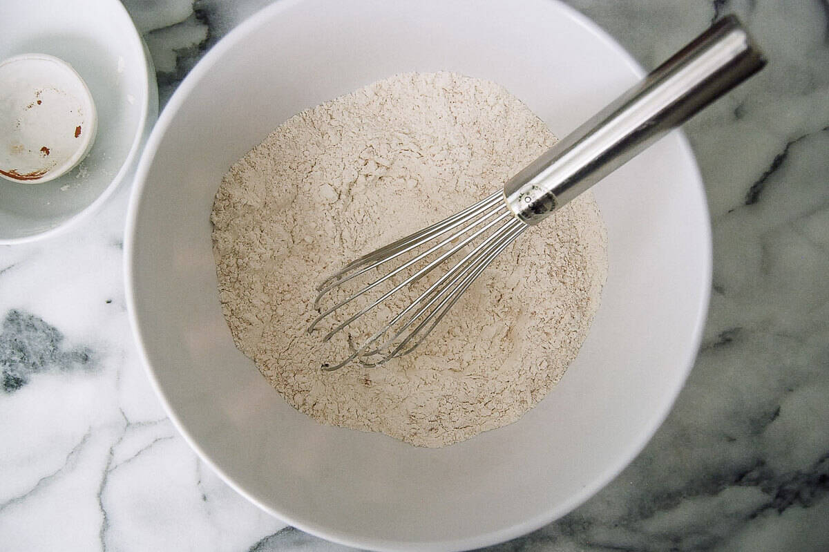 A metal whisk rests in a white bowl filled with flour, sitting on a marble countertop.