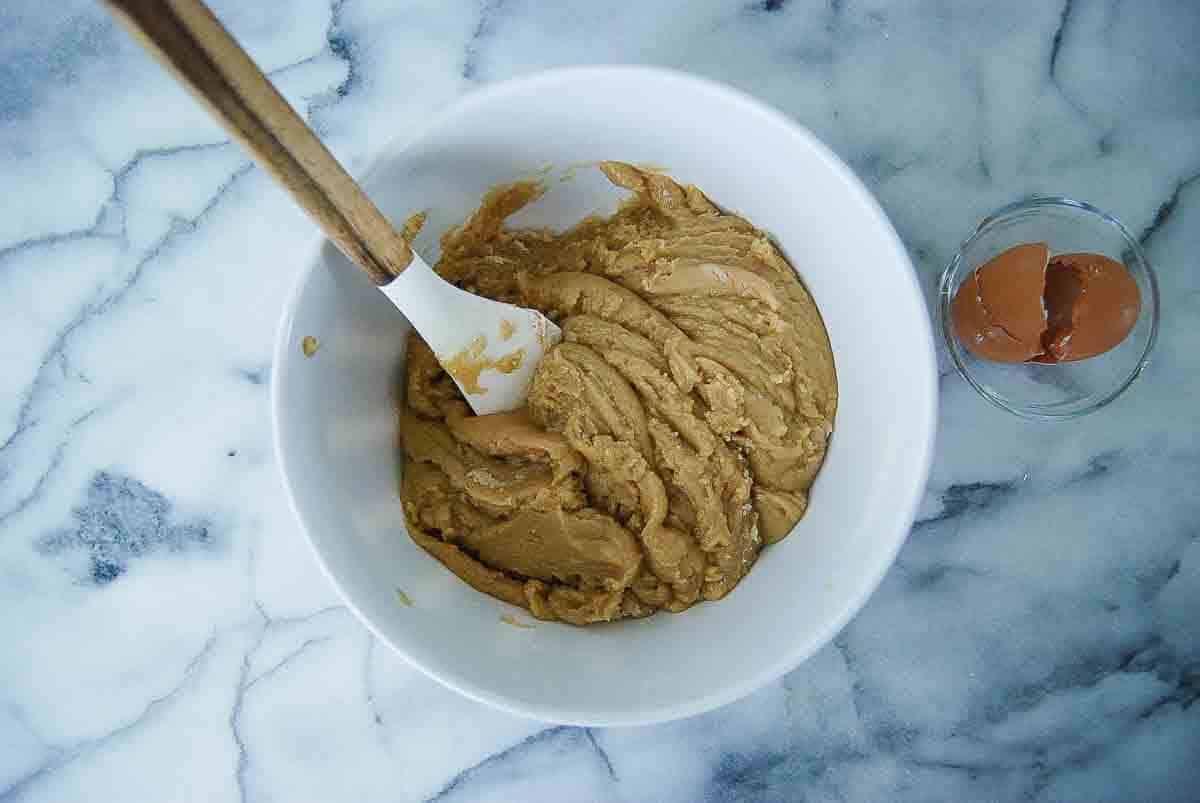 A white bowl with browned butter cookie dough being mixed by a spatula sits on a marble surface.