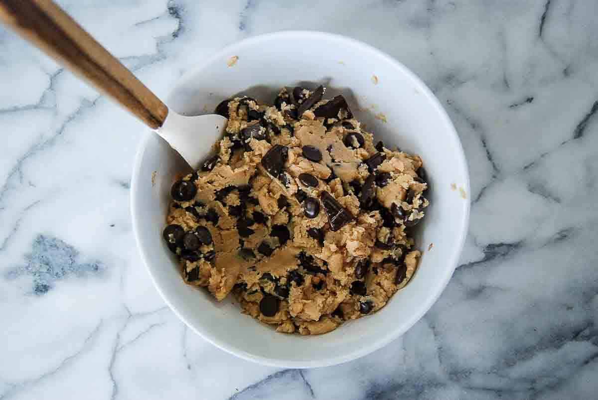 A white bowl filled with chocolate chip cookie dough sits on a marble countertop. A spatula with a wooden handle rests inside the bowl.